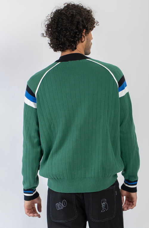 Jersey de punto verde con media cremallera, cuello alto y detalles deportivos en blanco, negro y azul.