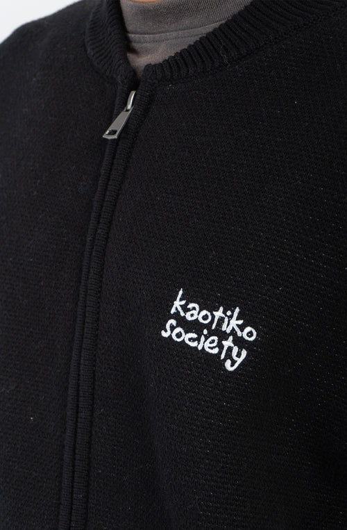 Jersey unisex de punto negro con cremallera, bordado Kaotiko Society en el pecho y gráfico jacquard de flores azules con mensaje Bad Romance en la espalda.