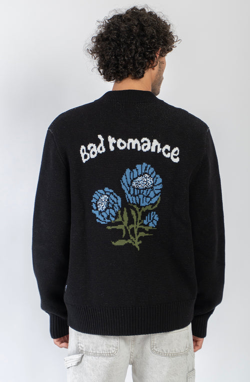 Jersey unisex de punto negro con cremallera, bordado Kaotiko Society en el pecho y gráfico jacquard de flores azules con mensaje Bad Romance en la espalda.