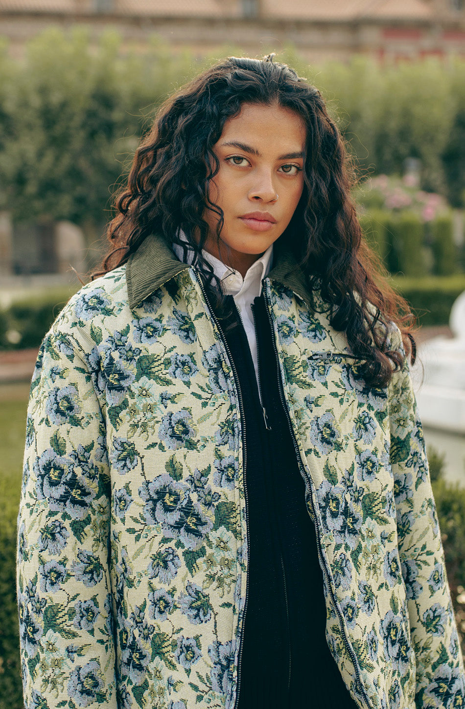 Chaqueta unisex de tejido tapestry con estampado floral en tonos verde, azul y crema, cuello de pana en contraste, cierre de cremallera y bolsillo de pecho con cremallera.